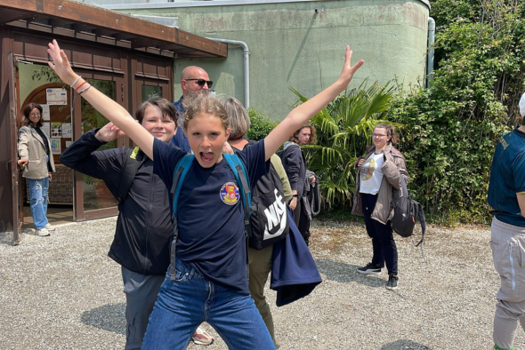 Orphelins d'Orpéopolis à l'extérieur devant un bâtiment. Au centre, un enfant vêtu d'un t-shirt bleu et d'un sac à dos adopte une pose énergique les bras levés en V, souriant à la caméra. D'autres enfants et adultes discutent ou marchent autour, certains