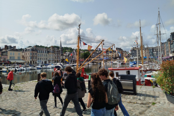 Orphelins d'Orpéopolis le long d'un quai pavé dans un port pittoresque. Plusieurs bateaux colorés, décorés de fanions, sont amarrés dans l'eau. À l'arrière-plan, on aperçoit des bâtiments anciens et des terrasses de cafés sous un ciel partiellement nuageu
