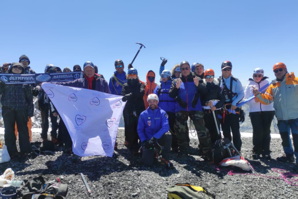 Orphelins d'Orphéopolis posant avec une bannière blanche, équipement d’alpinisme au sol, montagnes en arrière-plan.