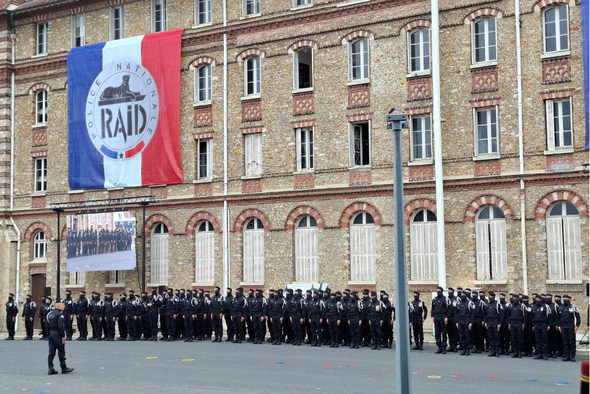 Les agents du RAID au garde à vous devant leurs emblèmes certains avec des drapeaux noirs