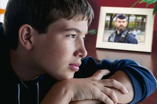 un jeune garçon est adossé sur un bureau le regard dans le vide. A côté de lui, une photo est encadrée d'un homme en uniforme de police. 